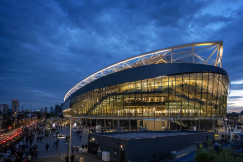 Tottenham Hotspur Stadium exterior glowing at dusk with crowds on the concourse